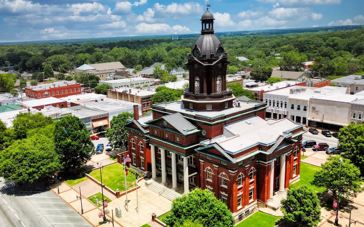 Aerial view of a historic red brick courthouse with a cupola tower surrounded by trees and buildings in a small town